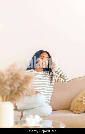 Thoughtful African-American woman sitting on couch in living room and dreaming, selective focus Stock Photo