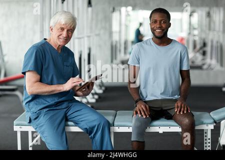 Portrait of senior rehabilitation therapist smiling at camera while consulting young man in clinic Stock Photo