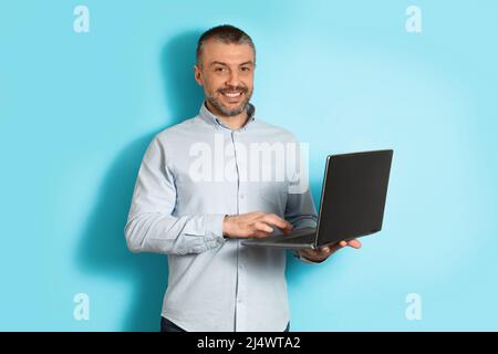 Middle Aged Man Using Laptop Computer Standing Over Blue Background Stock Photo