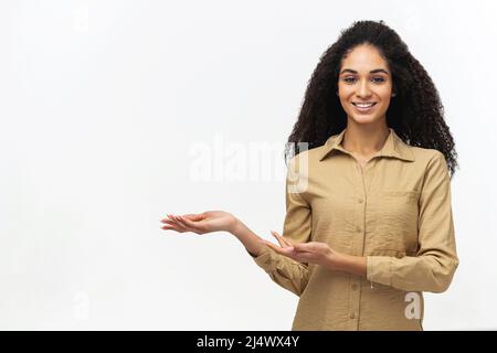 Charming multiracial woman with Afro hairstyle indicating palms at empty space isolated on white background, smiling female student advertising amazing novelty Stock Photo