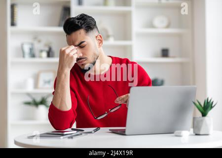 Tired attractive millennial arabic man with beard in red clothes takes off glasses at table with laptop in home Stock Photo