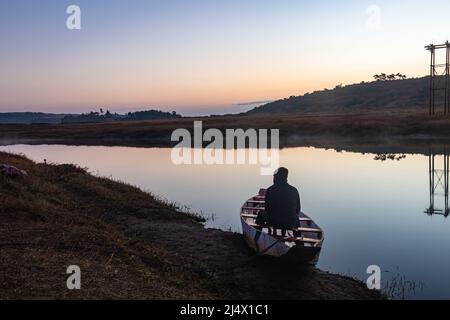 man sitting at traditional wood boat at calm lake with dramatic colorful sky reflection at morning Stock Photo