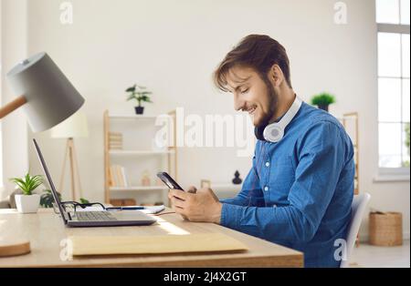 Happy young man using mobile phone while sitting at work desk with laptop at home Stock Photo