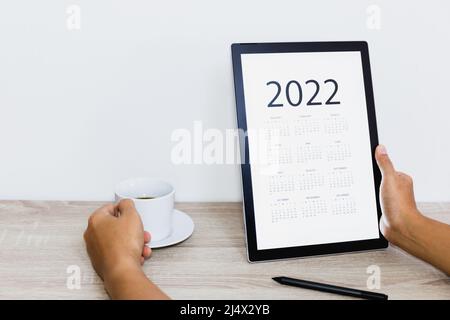 Hand holding coffee cup while looking calendar on digital tablet. Stock Photo