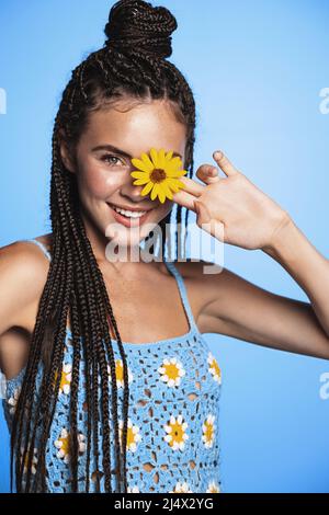 Beauty portrait of happy young woman enjoying vacation, posing with flower near her tanned face, standing in summer dress against blue background Stock Photo
