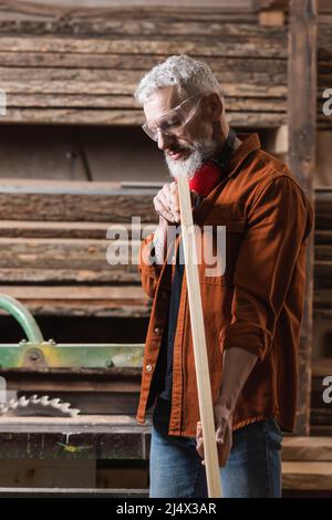 grey haired furniture designer in goggles checking wooden plank in woodwork atelier Stock Photo