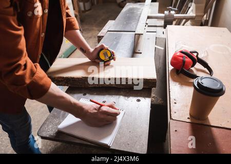 cropped view of carpenter writing in notebook near coffee to go on workbench Stock Photo