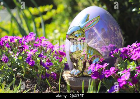 Golden easter bunny and egg sitting between lilac flowers in green garden. Stock Photo
