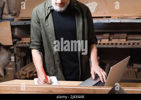 cropped view of furniture designer writing in notebook near laptop on workbench Stock Photo