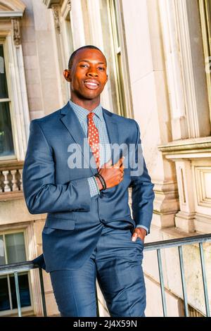 Successful businessman. Dressing in blue suit, tie, putting his hand on chest, a young black guy sitting on railing in vintage style office building, Stock Photo