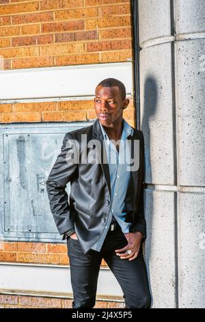 Man Waiting on Street.  Wearing black blazer, unbuttoned, striped under shirt, short haircut, a young black worker standing against brick wall and col Stock Photo