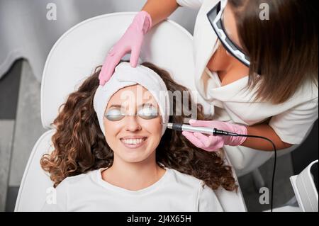 Top view of smiling young woman in silver protective goggles receiving laser skincare treatment in beauty salon. Cosmetologist using diode laser device while performing facial skincare procedure. Stock Photo