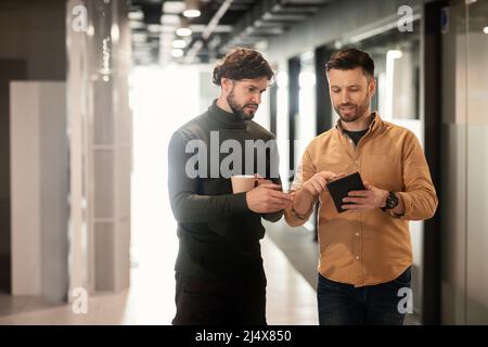 Two focused colleagues walking in company hall, using tablet pc, discussing business startup project at office, empty space Stock Photo