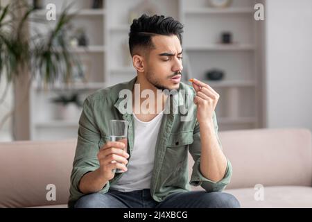Sad young arab man with beard in casual sits on sofa with glass of water and looks at pill in hand in living room Stock Photo