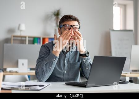 Mature male employee rubbing tired irritated eyes, working with laptop too much, exhausted from online job at office Stock Photo