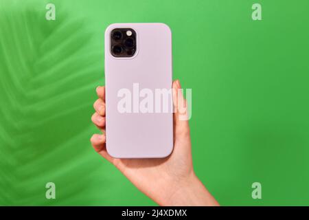 Female hand holding white smartphone in soft silicone cover back view . Phone case mock up isolated on green background shadow from a plant Stock Photo