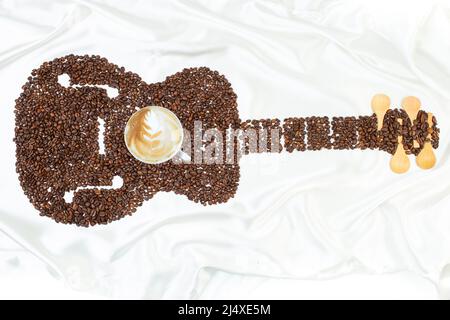 Guitar shape made from coffee beans with a cup of cappuccino in the middle on white satin fabric, music cafe album cover Stock Photo