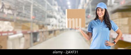 Young delivery woman in work clothes in front of a huge warehouse holding cardboard box for delivery to the customer. Stock Photo