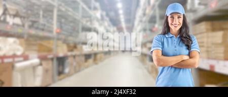 Young delivery woman in work clothes in front of a huge warehouse with crossed arms looking straight into the camera. Stock Photo