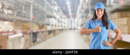 Young delivery woman in work clothes in front of a huge warehouse pointing finger up. Stock Photo