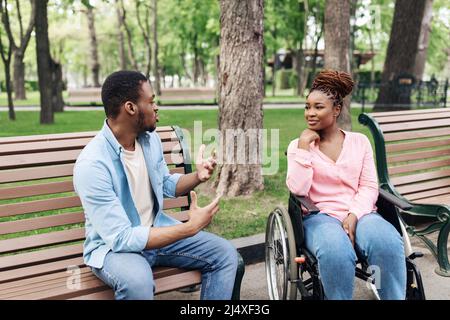 Young African American man sitting on bench, speaking to his female friend with physical disability outdoors Stock Photo