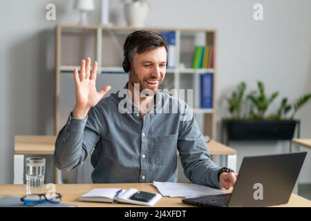 Happy mature caucasian businessman in headphones working on laptop and waving his hand to webcam, sitting in office Stock Photo