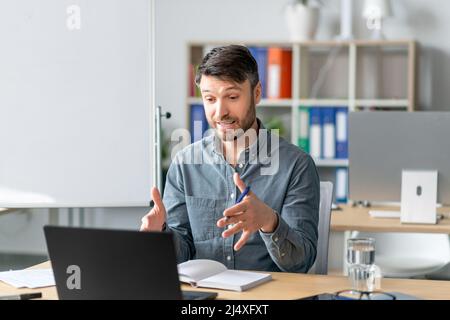 Mature male teacher looking at laptop and gesturing, sitting in office interior, explaining rules to student remotely Stock Photo