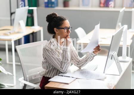 Paperwork concept. Focused female office worker reading paper or financial document and using computer in office Stock Photo