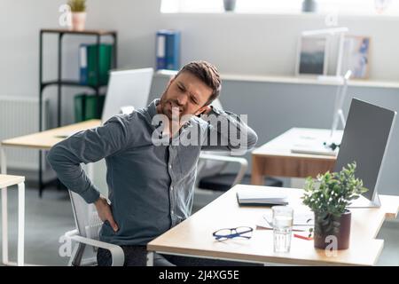 Mature businessman massaging aching neck and back, suffering from pain after computer work, sitting at workplace Stock Photo