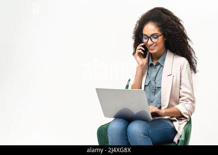 Confident African-American woman in smart casual wear using laptop and talking on the smartphone isolated on white background, female office employee typing and connecting with customers Stock Photo