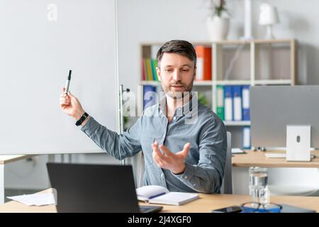 Male tutor conducting remote lesson near empty blackboard, using laptop for distance education, sitting in office Stock Photo