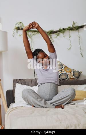 African pregnant woman sitting on bed and stretching while waking up in the morning Stock Photo