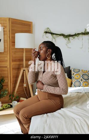 African pregnant woman sitting on bed and doing inhalation, she suffering from asthma during pregnancy Stock Photo