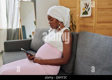 African pregnant muslim woman using her mobile phone for online reading while resting on sofa at home Stock Photo