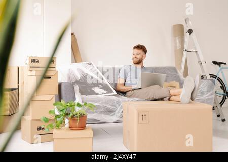 A relaxed young man sitting on the sofa using his laptop while smiling Stock Photo