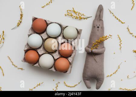 Funny handmade bunny rabbit toy with colorful chicken eggs and yellow mimosa flowers. Easter gift concept.  Stock Photo