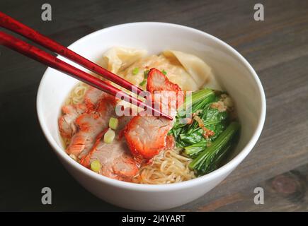 Bowl of Mouthwatering Egg Noodle and Wonton Dumpling Soup with Roasted Pork on Wooden Table Stock Photo