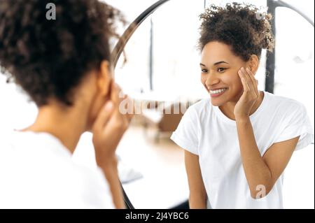 Lovely happy African American curly-haired girl in a white basic t-shirt, stands in front of a mirror in the morning, admires herself, smiles, starts the day well, good mood, looks at her face Stock Photo