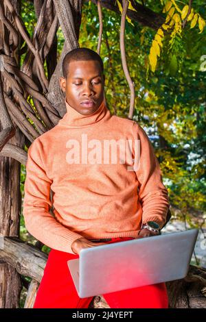 Man Working Outside. Dressing in light orange sweater with high collar, red pants, wearing a wristwatch, a young handsome black guy is sitting on wood Stock Photo