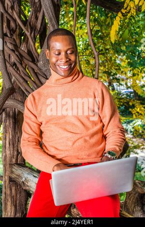 Man Working Outside. Dressing in light orange sweater with high collar, red pants, wearing a wristwatch, a young black guy is sitting on wood area, lo Stock Photo