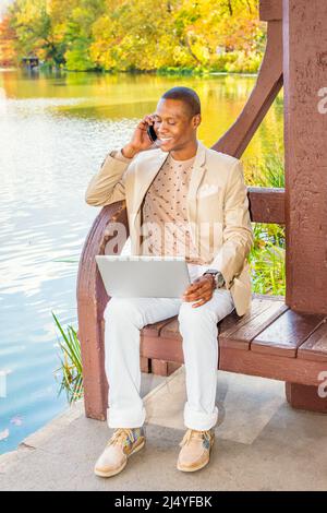 Business Man Working. Dressing in cream blazer, collarless sweater, white pants, sneakers, a young black guy is sitting by a lake, smiling, talking on Stock Photo