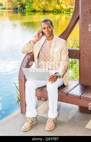 Business Man Working Outside. Dressing in cream blazer, collarless sweater, white pants, sneakers, a young black guy is sitting by a lake, talking on Stock Photo