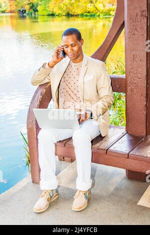 Business Man Working Outside. Dressing in cream blazer, collarless sweater, white pants, sneakers, a young black guy is sitting by a lake, talking on Stock Photo