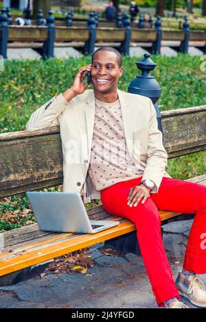 Man Working Outside. Dressing in cream blazer, collarless sweater, red pants, sneakers, a young black guy is sitting on the bench, smiling, making pho Stock Photo
