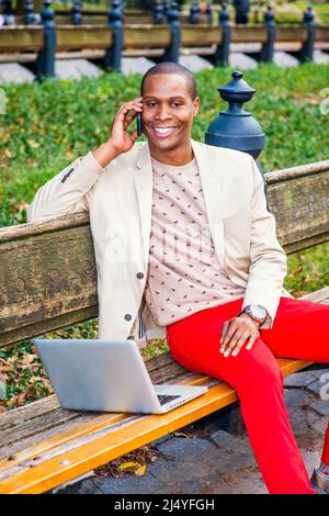 Man Working Outside. Dressing in cream blazer, collarless sweater, red pants, sneakers, a young black guy is sitting on the bench, smiling, making pho Stock Photo