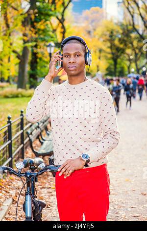 Man Listening Music. Dressing in collarless sweater, red pants, wearing headphone, a young black guy is standing on street, holding a bike, a hand tou Stock Photo