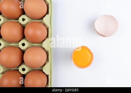 Pack of brown eggs with one broken open egg with raw yolk and shell on white background. Stock Photo
