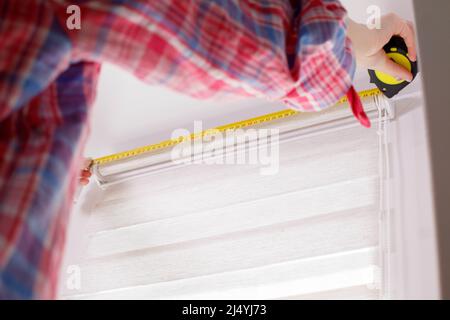 A handywoman with a tape measure installs white roller blinds indoors. Shutters day and night Stock Photo