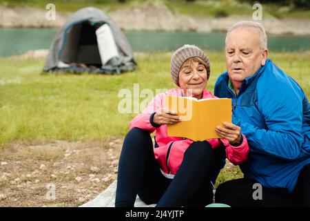 Senior couple having fun reading a book while camping in mountain lake - Focus on woman face Stock Photo
