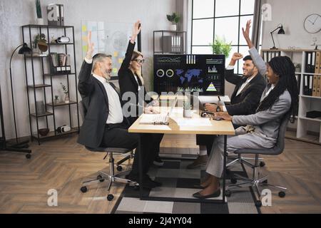 Smiling diverse cheerful multiracial businesspeople sit at office desk, celebrating successful project, gesturing, laughing, clapping hands and sharing business ideas together. Stock Photo